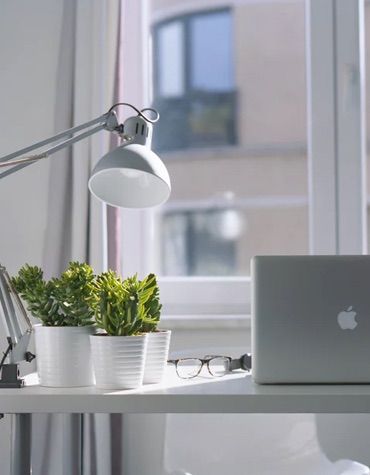 macbook on a desk by an open window with a lamp, three plants, and glasses