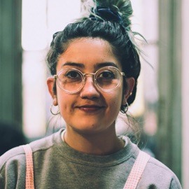woman with brown hair in a top knot wearing a grey sweatshirt with pink overalls overtop and glasses